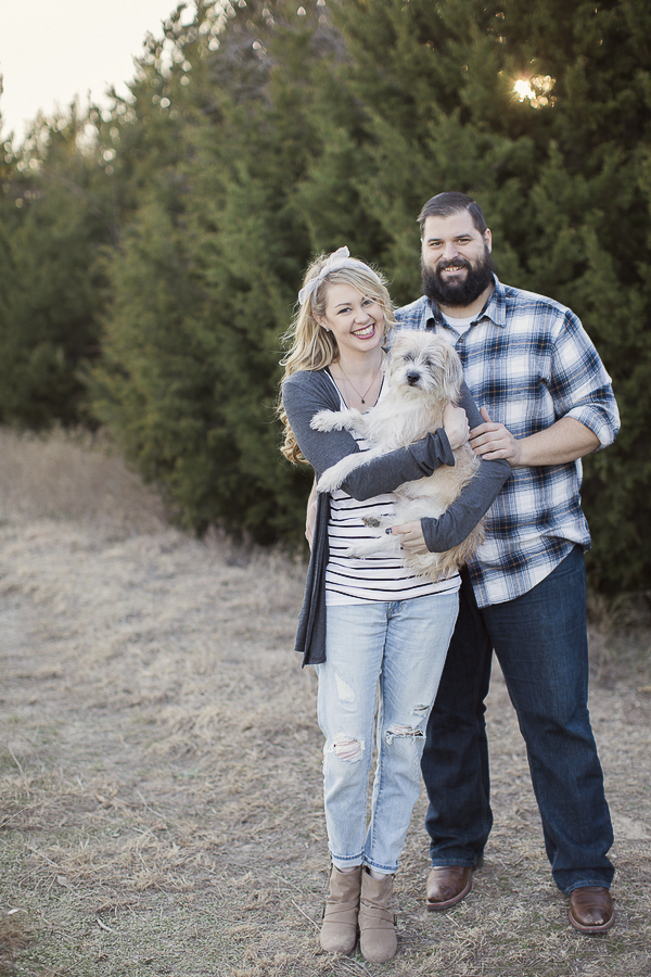 woman holding terrier mix, man, engaged couple in front of pines