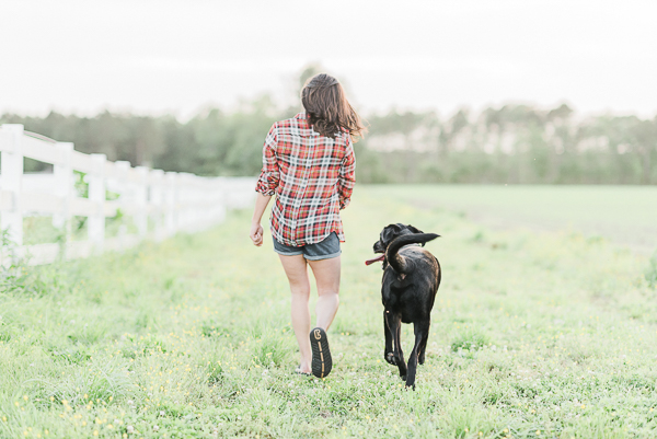 black lab and woman in plaid shirt and jean shorts walking away through a field