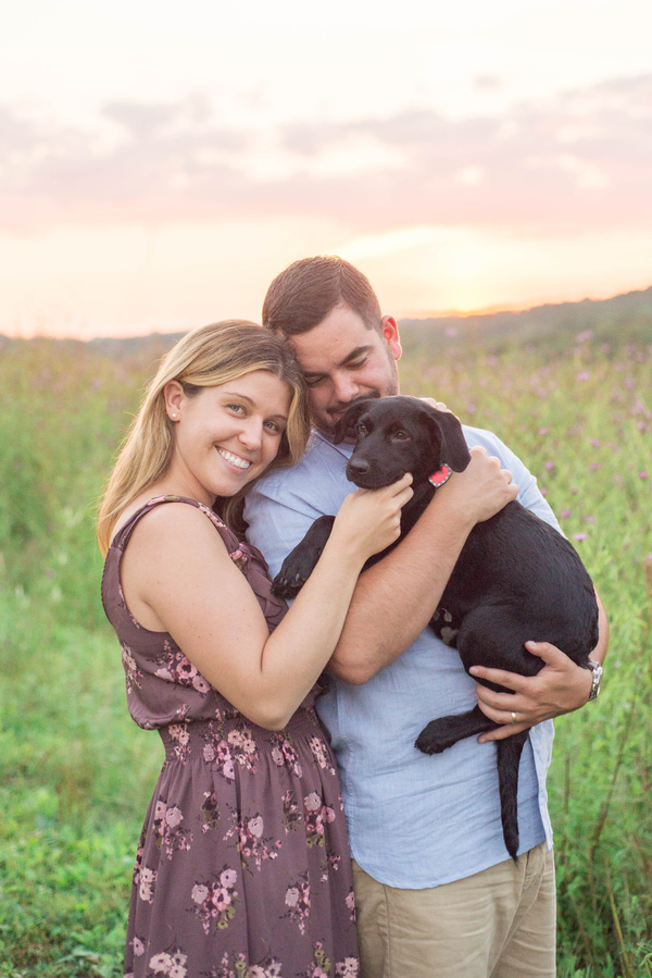 woman and man standing in field holding adorable black lab mix puppy