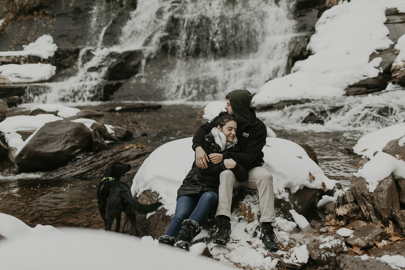 dog looking at waterfall while couple snuggles on boulder, winter engagement session | ©Belle La Vie Images 