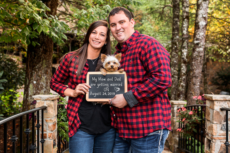couple wearing buffalo plaid shirts, holding save the date sign with Yorkie | ©Cariad Photography | dog-friendly engagement session, Clayton, GA