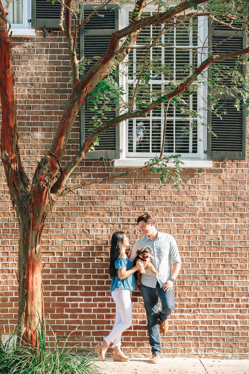 couple holding small dog and leaning against brick building, dog-friendly engagement poses, ©Charleston Photo Art