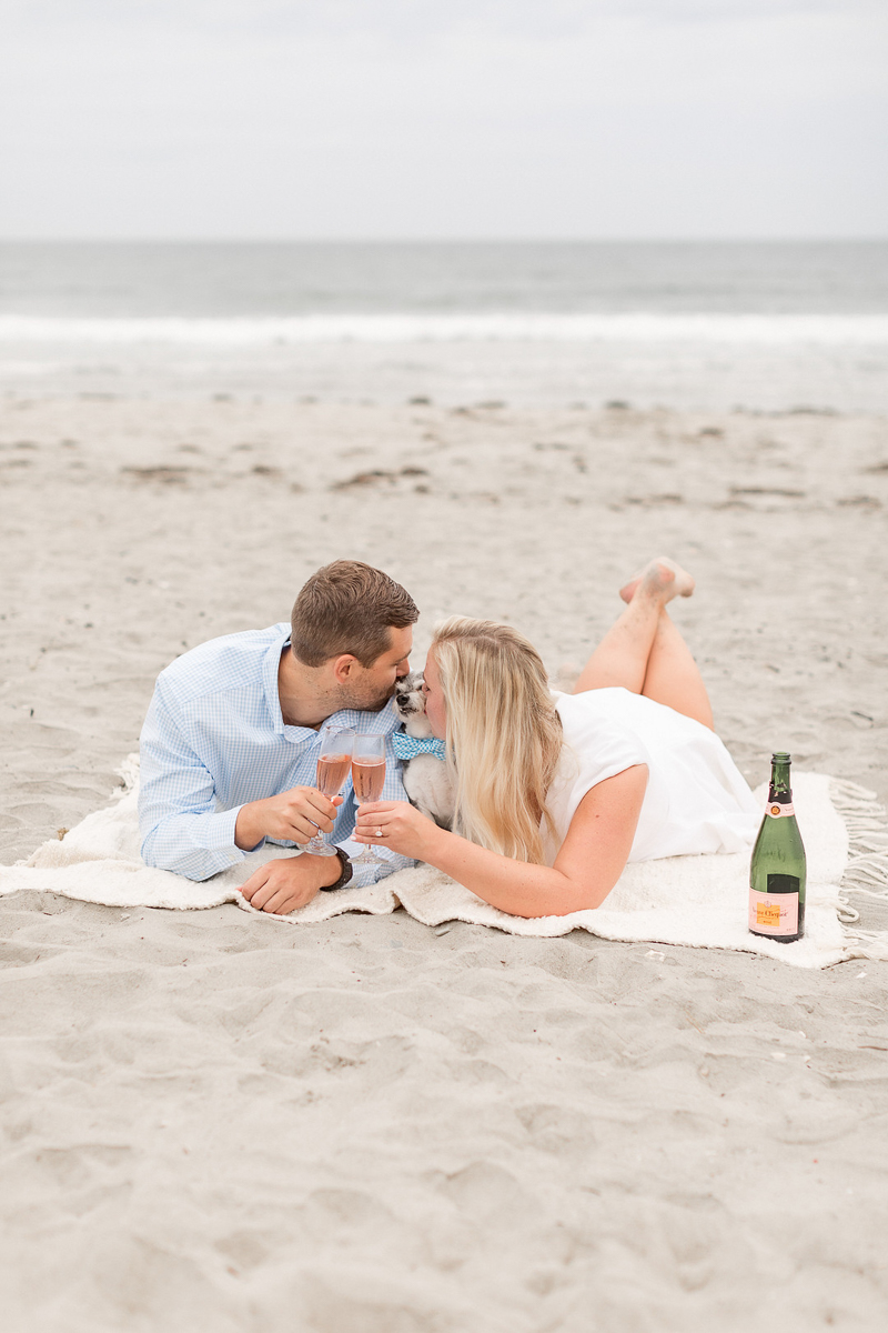 couple on blanket kissing their small dog and holding champagne glasses, beach engagement portraits | ©Coli Michael Photography 