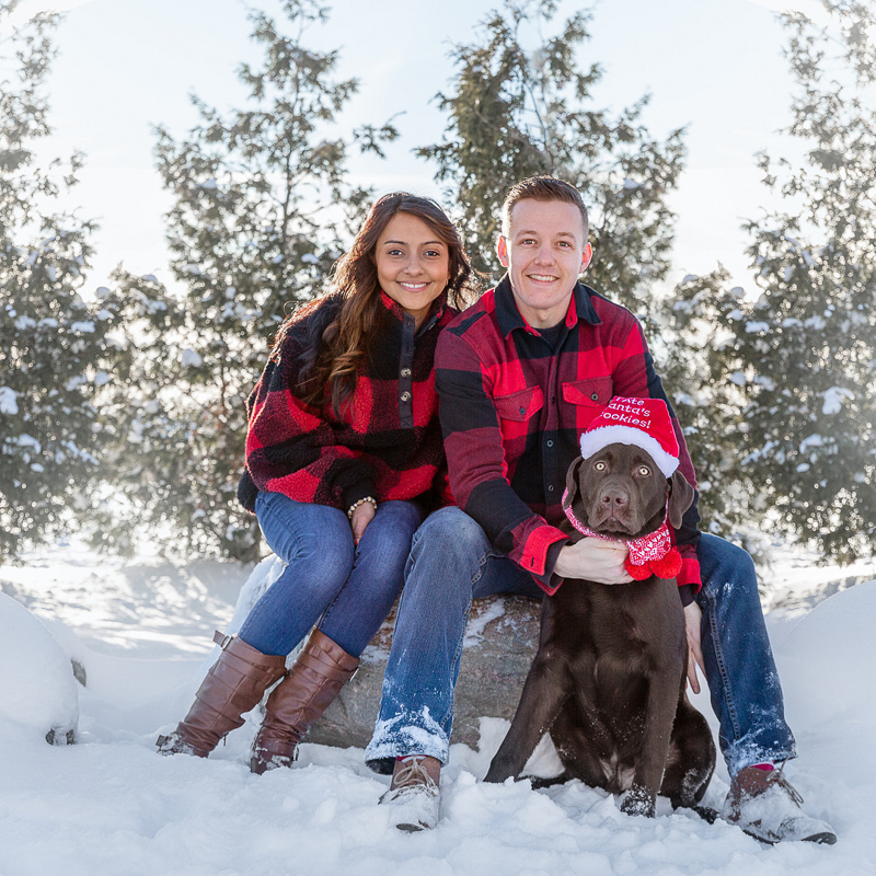 couple wearing buffalo plaid jackets sitting on a rock, Chocolate Lab wearing hat that says "I ate Santa's cookies" | © Terri J Photography