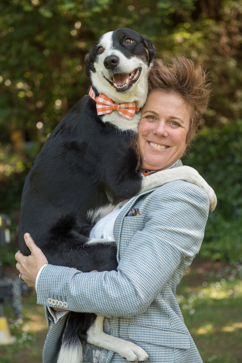 bride wearing suit holding black and white dog in orange gingham bow tie, Rustically Romantic Photography by Darby Johnson