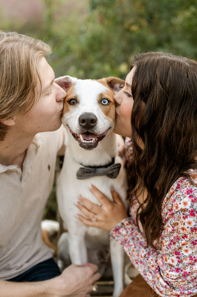 couple kissing white and brown dog wearing bow tie, dog-friendly engagement | ©The Ramseys Photography