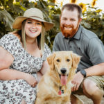 woman wearing straw hat and dress kneeling next to man in short sleeved short and shorts with their dog in a sunflower field | ©Ashley Kalbus Photography, Green Bay, WI Photography